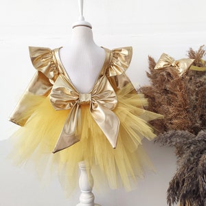 Puede incluir: Un vestido de tul amarillo con un lazo dorado met&aacute;lico y mangas con volantes. El vestido est&aacute; en un maniqu&iacute; blanco.