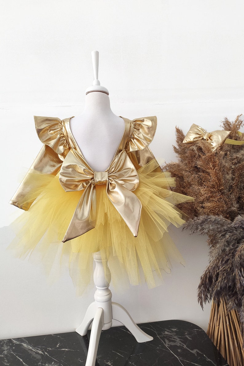 Puede incluir: Un vestido de tul amarillo con un lazo dorado met&aacute;lico y mangas con volantes. El vestido est&aacute; en un maniqu&iacute; blanco.