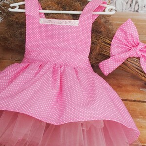 Può includere: Un vestito rosa a pois bianchi con gonna in tulle e fiocco abbinato. L'abito ha uno scollo quadrato e un bordo in pizzo bianco. Il fiocco è grande e presenta lo stesso motivo a pois dell'abito. L'abito è appeso a un appendiabiti bianco.