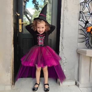 Könnte beinhalten: Ein junges Mädchen, das sich für Halloween als Hexe verkleidet hat. Sie trägt ein schwarzes und violettes Kleid mit einem Tüllrock und einen schwarzen Hexenhut.