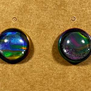 May include: Two iridescent stud earrings with a dark, multi-colored, swirling design. The earrings have a dark border and are set against a tan background. The earrings are round and appear to be made of glass or resin.