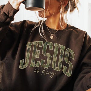 May include: A brown sweatshirt with the text "JESUS is King" in a camouflage design. The person is holding a black mug. The sweatshirt is long-sleeved and the person is wearing fuzzy slippers.