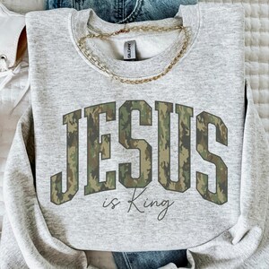 May include: A light gray sweatshirt with the text "JESUS is King" in a camouflage pattern. The sweatshirt is paired with blue jeans, white sneakers, and a gold chain necklace. The items are arranged on a white surface.