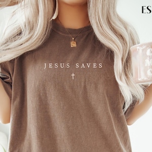 Puede incluir: Camiseta color espresso con el texto "JESUS SAVES" y una cruz. La camiseta es de corte holgado y tiene cuello redondo. La modelo lleva una goma para el pelo y sostiene una taza.