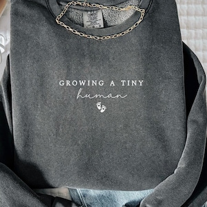 May include: A charcoal gray sweatshirt with the text "GROWING A TINY human" in white script, along with tiny foot icons. A gold chain necklace rests on the neckline. The sweatshirt is paired with blue jeans and white sneakers.