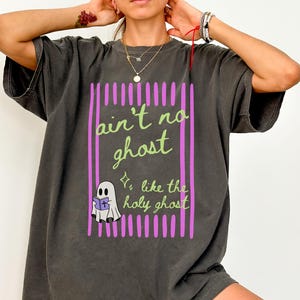May include: A charcoal gray short-sleeve t-shirt featuring a graphic print with vertical magenta stripes. The text reads "ain't no ghost like the holy ghost" in light green script, accompanied by a small white ghost illustration holding a purple book.