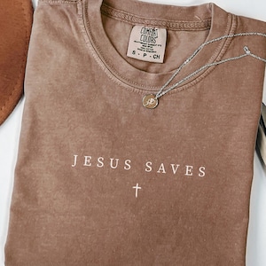 Puede incluir: Camiseta color espresso con el texto "JESUS SAVES" y una cruz. Un collar plateado con un colgante descansa sobre la camiseta. La camiseta est&aacute; doblada y tiene una etiqueta Comfort Colors. Un sombrero marr&oacute;n es visible en el fondo.