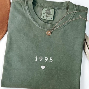 May include: A green t-shirt with the year 1995 and a heart printed on the front. The shirt is made by Comfort Colors.