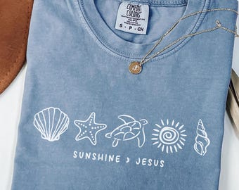 Könnte beinhalten: Ein kurzärmeliges T-Shirt in Jeansblau mit einem weißen Grafikdruck, der eine Muschel, einen Seestern, eine Meeresschildkröte, eine Sonne und eine Spiralschnecke zeigt. Unter den Symbolen steht der Text SUNSHINE & JESUS auf dem Stoff.