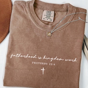 May include: Espresso-colored t-shirt with the text "fatherhood is kingdom work" and "PROVERBS 22:6" in white. A silver necklace with a pendant and a cross graphic are also visible. The shirt is a Comfort Colors brand.