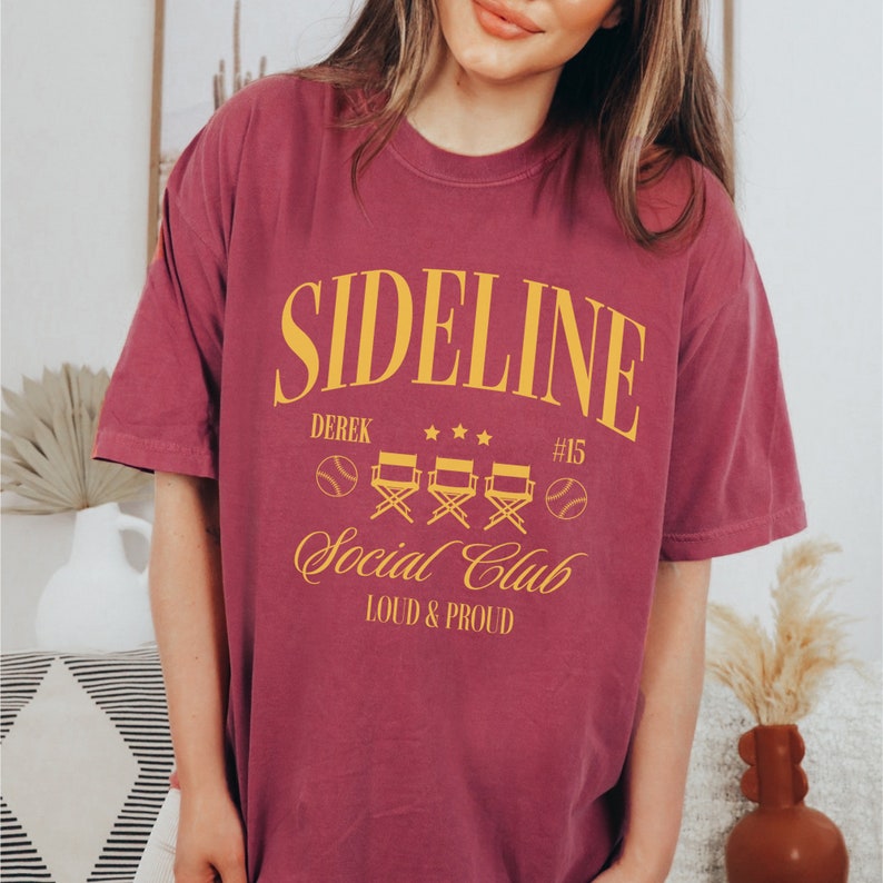 Puede incluir: Una camiseta color granate con un gr&aacute;fico amarillo que dice "Sideline Social Club" con el texto "Derek #15" y "Loud & Proud" debajo. El gr&aacute;fico tambi&eacute;n incluye una pelota de b&eacute;isbol y cuatro sillas de director.