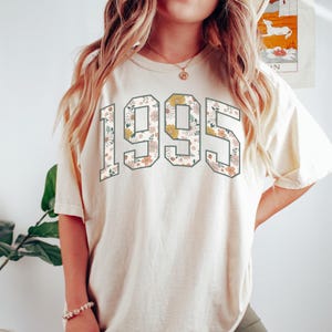 May include: A cream colored t-shirt with a floral print of the year 1995.