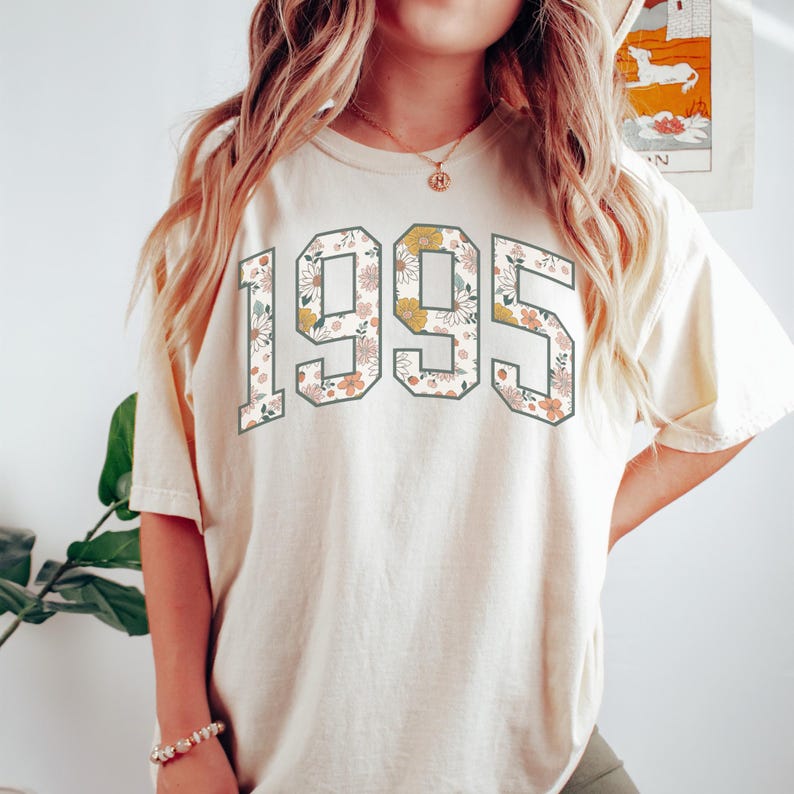 May include: A cream colored t-shirt with a floral print of the year 1995.