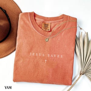 Puede incluir: Una camiseta de color coral con las palabras "JESUS SAVES" y una cruz blanca. Tambi&eacute;n se ven un collar dorado con un colgante y un sombrero marr&oacute;n. La camiseta es de Comfort Colors.