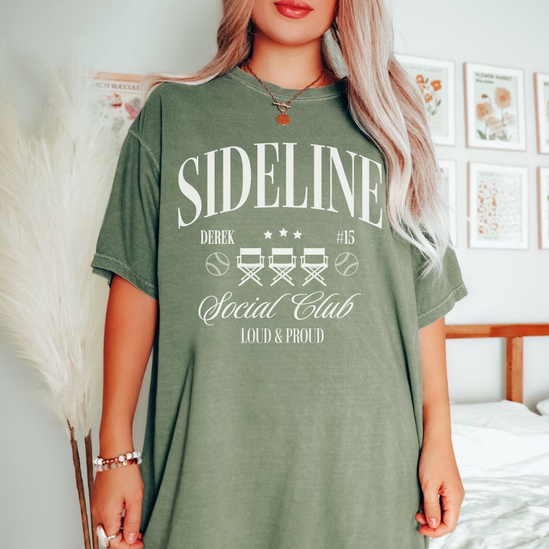 Puede incluir: Una camiseta verde con el texto "SIDE LINE" en blanco, con las palabras "Derek", "Social Club" y "Loud & Proud" debajo. Hay cuatro sillas de director blancas y dos pelotas de b&eacute;isbol blancas en la camiseta.