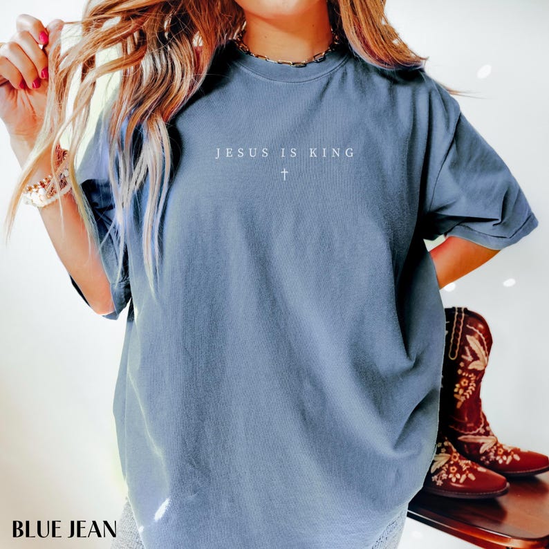 May include: Blue jean colored t-shirt with the text "JESUS IS KING" and a cross symbol. The shirt is a solid color and has short sleeves. The words and cross are in white. The bottom left corner of the image has the words "BLUE JEAN".