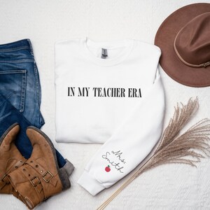 Pu&ograve; includere: Felpa bianca con la scritta "In My Teacher Era" sul davanti e "Mrs. Smith" con una mela rossa sulla manica sinistra.