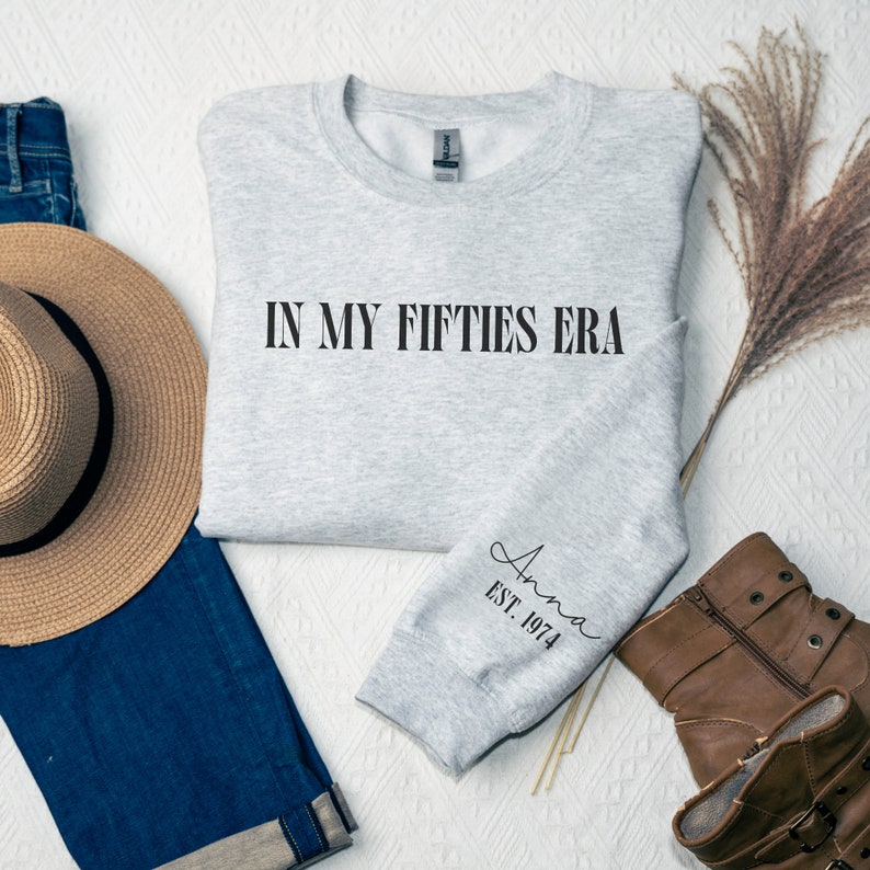 May include: A gray sweatshirt with the text "IN MY FIFTIES ERA" printed on the front and "Anna EST. 1974" on the sleeve.