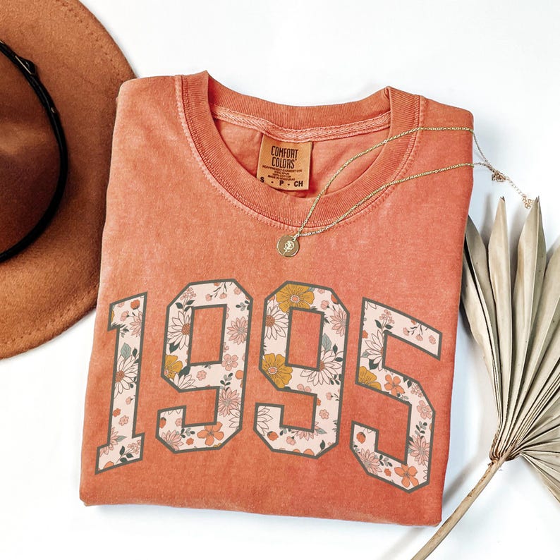 May include: A coral-colored t-shirt with a floral print design of the year 1995. The shirt has a distressed look and is made of a soft, comfortable fabric.
