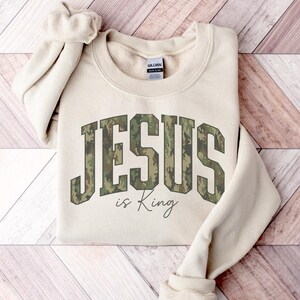 May include: A beige sweatshirt with the words "JESUS is King" printed on the front. The word "JESUS" is in large, block letters with a camouflage pattern. The words "is King" are in a cursive font.