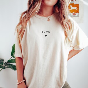 May include: A white t-shirt with the year 1995 printed on the front, along with a small black heart.