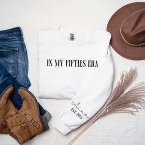 May include: White sweatshirt with the text "IN MY FIFTIES ERA" printed on the front and the text "Anna EST. 1974" printed on the left sleeve.