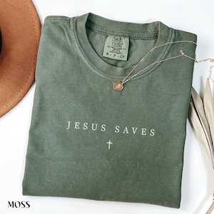 Puede incluir: Una camiseta verde musgo con las palabras "JESUS SAVES" y una cruz blanca. La camiseta tiene cuello redondo y mangas cortas. Un collar dorado con un colgante descansa sobre la camiseta. La camiseta est&aacute; doblada.