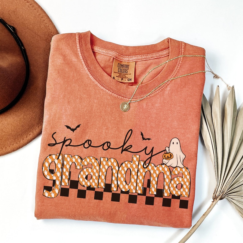 Op de afbeelding: Een verbrand oranje T-shirt met de tekst "spooky grandma" in zwart en wit. De tekst is omringd door een patroon van oranje en witte geesten. Rechts van de tekst staat een klein wit spook met een pompoen.