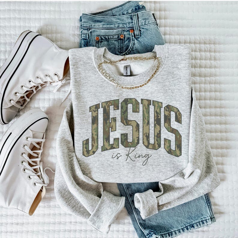 May include: A light gray sweatshirt with the text "JESUS is King" in a camouflage pattern. The sweatshirt is paired with blue jeans, white sneakers, and a gold chain necklace. The items are arranged on a white surface.