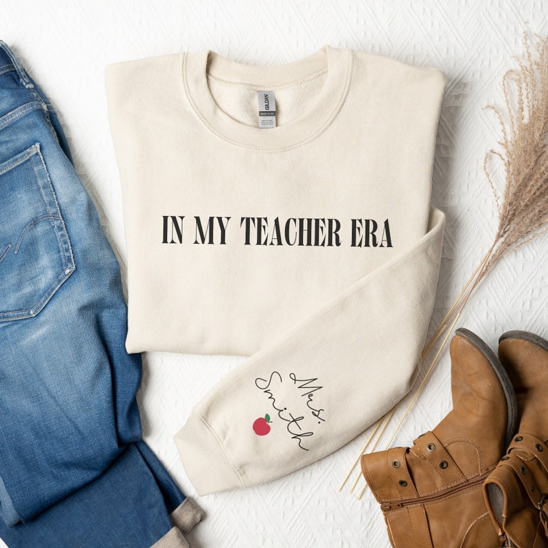 Pu&ograve; includere: Una felpa beige con la scritta "IN MY TEACHER ERA" stampata sul davanti e "Mrs. Smith" con una mela rossa sulla manica sinistra.