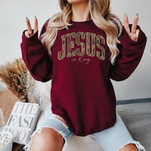 May include: Burgundy sweatshirt with the text "JESUS is King" in a camouflage design. The sweatshirt has long sleeves and a crew neck. The text is in a bold, collegiate-style font. The words "is King" are in a smaller, cursive font.