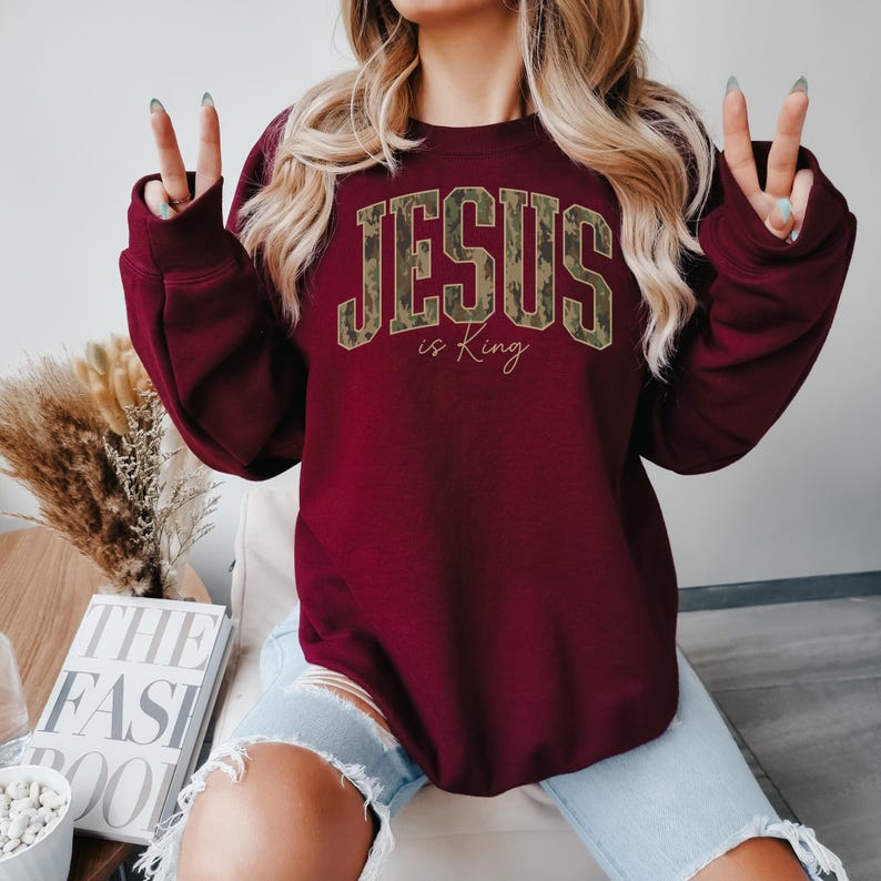 May include: Burgundy sweatshirt with the text "JESUS is King" in a camouflage design. The sweatshirt has long sleeves and a crew neck. The text is in a bold, collegiate-style font. The words "is King" are in a smaller, cursive font.