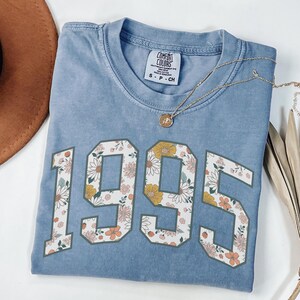 May include: A light blue t-shirt with a floral design in the numbers "1995".