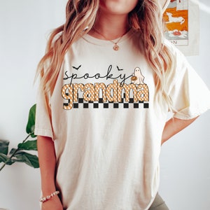 Op de afbeelding: Een wit T-shirt met de tekst "Spooky Grandma" in zwart en oranje. De tekst is in een geruit patroon met een spook dat een pompoen vasthoudt aan de rechterkant van de tekst.