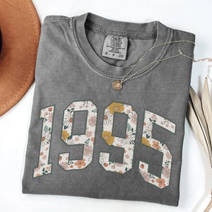 May include: A gray t-shirt with a vintage-style floral print of the year 1995. The shirt has a distressed look and is made of a soft, comfortable fabric.