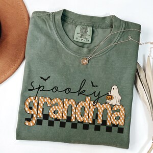 Op de afbeelding: Een groen T-shirt met de tekst "spooky grandma" in oranje en witte letters met een spook dat een pompoen vasthoudt. De letters zijn bedekt met een patroon van kleine witte spookjes.