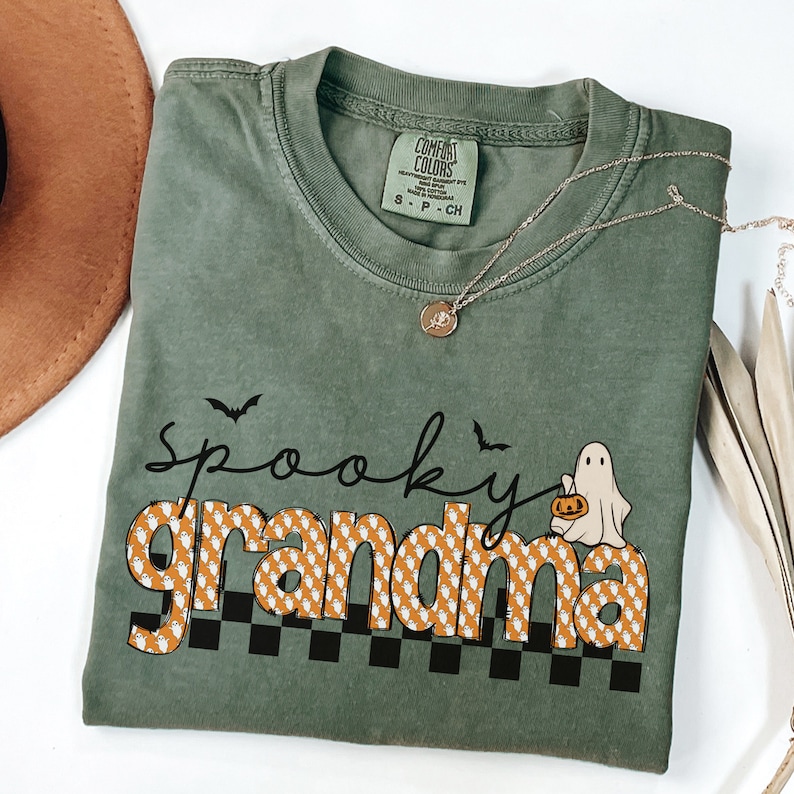 Op de afbeelding: Een groen T-shirt met de tekst "spooky grandma" in oranje en witte letters met een spook dat een pompoen vasthoudt. De letters zijn bedekt met een patroon van kleine witte spookjes.