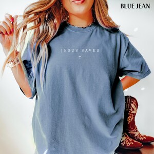 Puede incluir: Camiseta azul vaquero con el texto "JESUS SAVES" y una cruz. La camiseta tiene mangas cortas y un corte holgado. La imagen tambi&eacute;n muestra un par de botas de vaquero rojas.