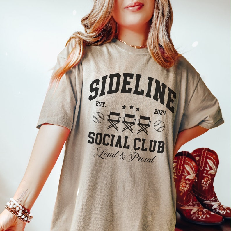Op de afbeelding: Een beige T-shirt met een zwart grafisch ontwerp met de tekst "Sideline Social Club Est. 2024 Loud & Proud". De grafiek bevat honkballen, regisseursstoelen en sterren.