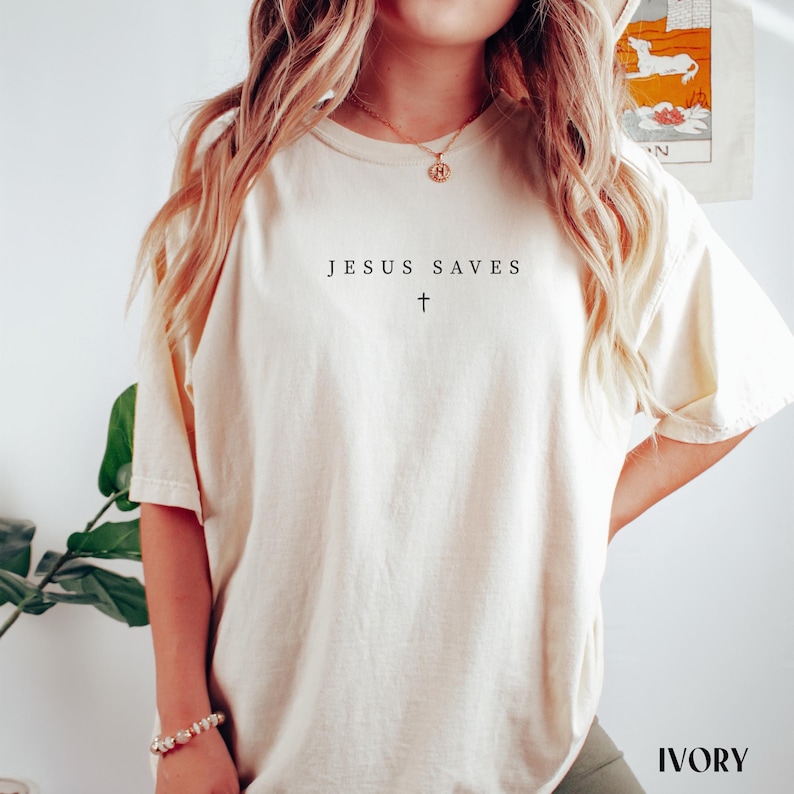 Puede incluir: Camiseta de color marfil con el texto "JESUS SAVES" y un s&iacute;mbolo de cruz impreso en negro. La camiseta es de corte holgado, con mangas cortas. La palabra "IVORY" est&aacute; impresa en una fuente serif en la esquina inferior derecha.