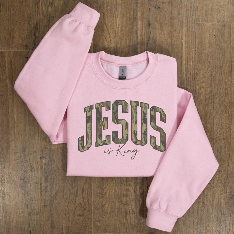 May include: A light pink sweatshirt with the words "JESUS is King" printed on the front. The word "JESUS" is in a camouflage pattern. The sweatshirt has long sleeves and a crew neck.