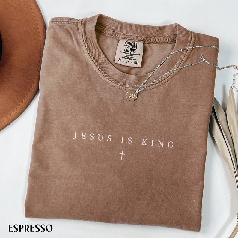 May include: Espresso-colored t-shirt with the text "JESUS IS KING" and a cross. The shirt has a crew neck and short sleeves. A silver necklace with a pendant is draped across the shirt. The shirt is folded on a white surface.