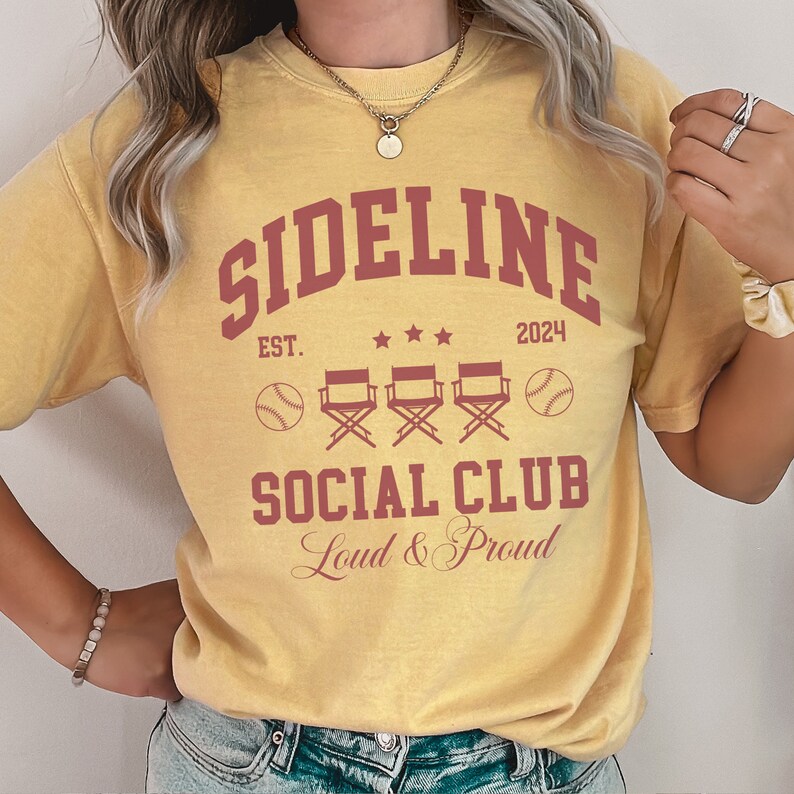 Op de afbeelding: Een geel T-shirt met de tekst "Sideline Social Club Est. 2024 Loud & Proud" in rood. Het ontwerp bevat ook drie regisseursstoelen en honkballen.
