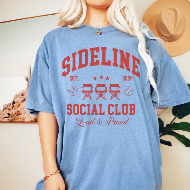 Op de afbeelding: Een lichtblauw t-shirt met een rode afbeelding waarop "Sideline Social Club" staat, met de tekst "Est. 2024" en "Loud & Proud" eronder. De afbeelding bevat ook drie rode regisseursstoelen en twee honkballen.