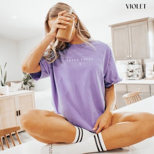 Puede incluir: Una camiseta violeta con el texto "JESUS SAVES" y una cruz. La camiseta se usa con calcetines blancos con rayas negras. Una persona sostiene un vaso transparente con una pajita.