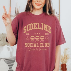 Op de afbeelding: Een maroon t-shirt met een vintage-stijl grafisch ontwerp dat "Sideline Social Club" zegt met de tekst "Est. 2024" en "Loud & Proud" eronder. De grafiek bevat ook honkballen en regisseursstoelen.