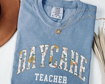 May include: A light blue t-shirt with the text "DAYCARE TEACHER" in a white arch with floral accents. The shirt is made of a soft, comfortable material.