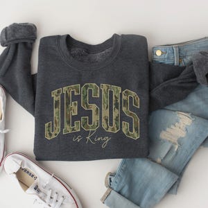 May include: A dark gray sweatshirt with the text "JESUS is King" in a camouflage design. The sweatshirt is paired with light blue jeans and white sneakers with red trim.