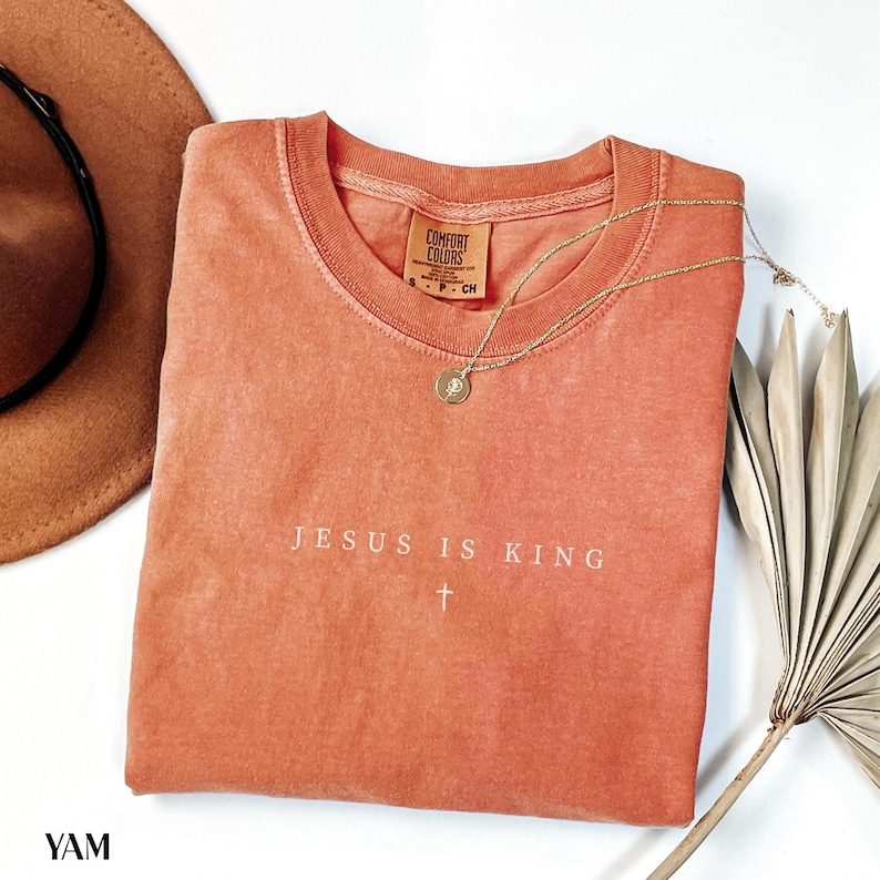 May include: A coral-colored t-shirt with the text "JESUS IS KING" and a cross graphic. A gold necklace with a pendant and a brown hat are next to the shirt. The shirt is folded on a white surface.