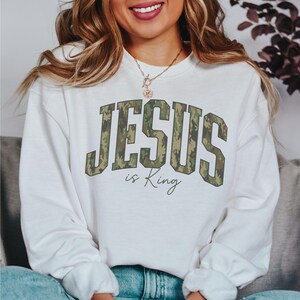 May include: White sweatshirt with the text "JESUS is King" in a camouflage design. The sweatshirt is a crew neck style and appears to be made of a soft material. The text is in a bold, collegiate-style font.